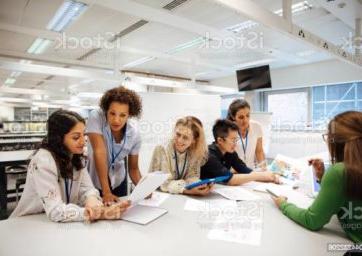 老师带着一群大学生，在实验室的教室里. 老师正在考虑一个学生的工作，心情轻松而积极. 其他同学正在互相讨论事情. 这是一个真实的教学场景，坦率的表达. 这是一个多民族的妇女群体. 背景中有一块写着数学公式的白板. All ladies are wearing id tags.